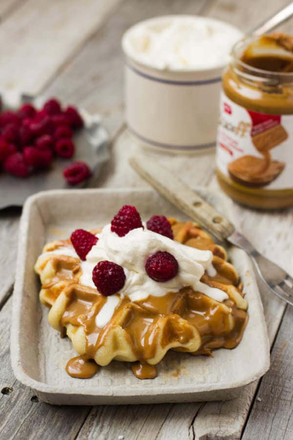 A liege waffle on a paper plate topped with cookie butter, whipped cream, and raspberries. 
