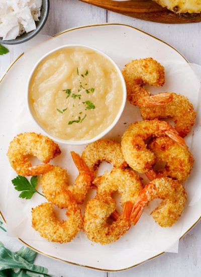 A small bowl of coconut shrimp sauce, that is a light yellow color, the bowl is on a plate surrounded by coconut shrimp.