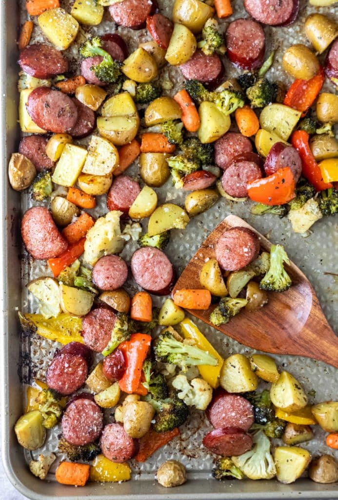 A baking pan topped with scattered slices of sausages, potatoes, carrots, broccoli, bell pepper, and cauliflower.