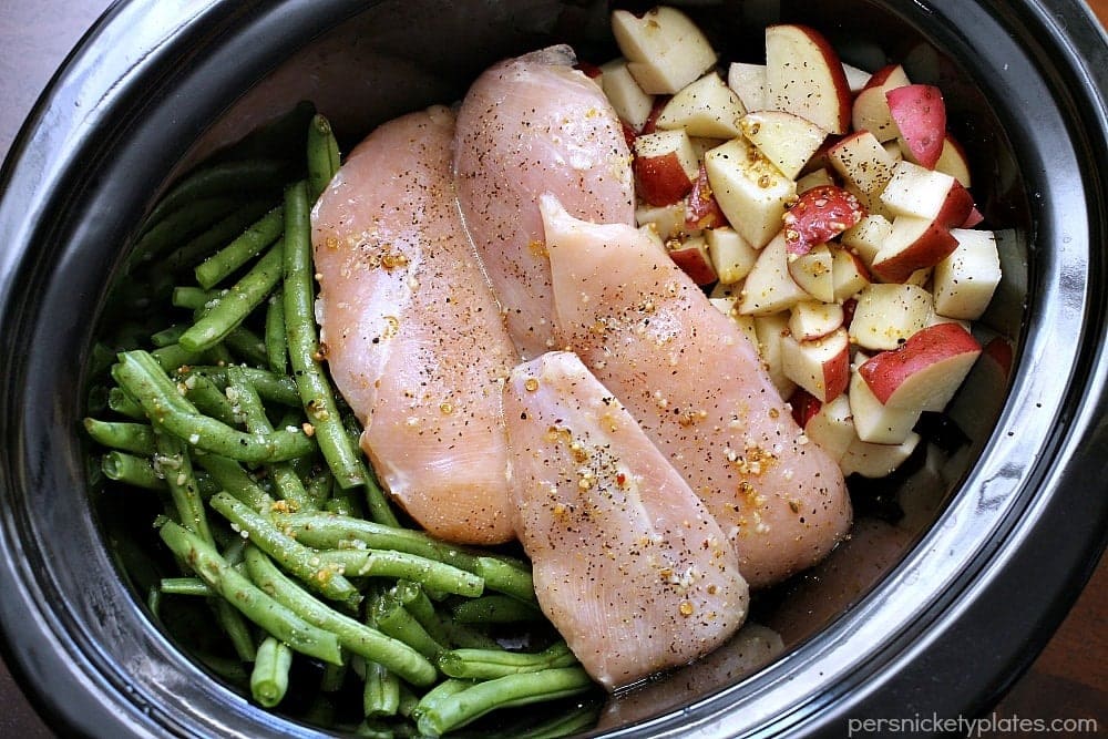 An overhead photo of an open slow cooker, full of green beans, raw chicken breasts, and diced red potatoes.