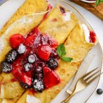 A plate of three rolled crepes topped with berries, and sauce on a plate.