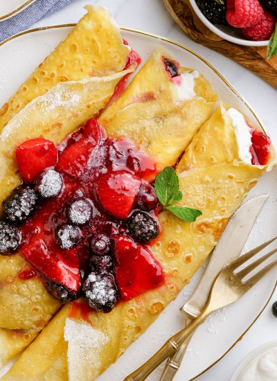 A plate of three rolled crepes topped with berries, and sauce on a plate.