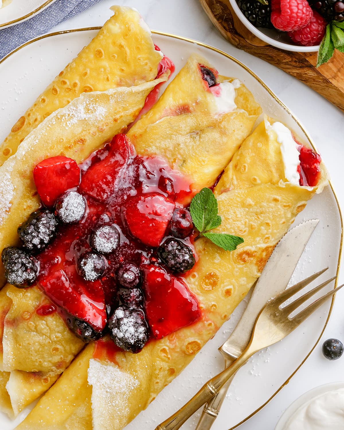 A plate of three rolled crepes topped with berries, and sauce on a plate.