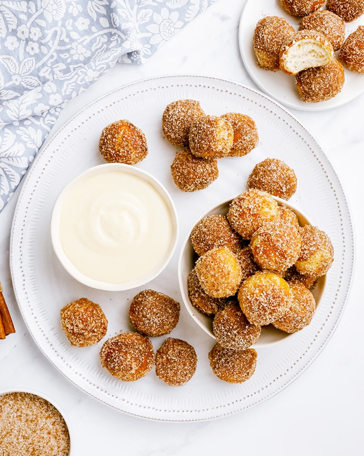 An overhead shot of a plate full of soft pretzel bites, shaped like little golf balls, and covered in cinnamon sugar. With a bowl of cream cheese dip next to them.