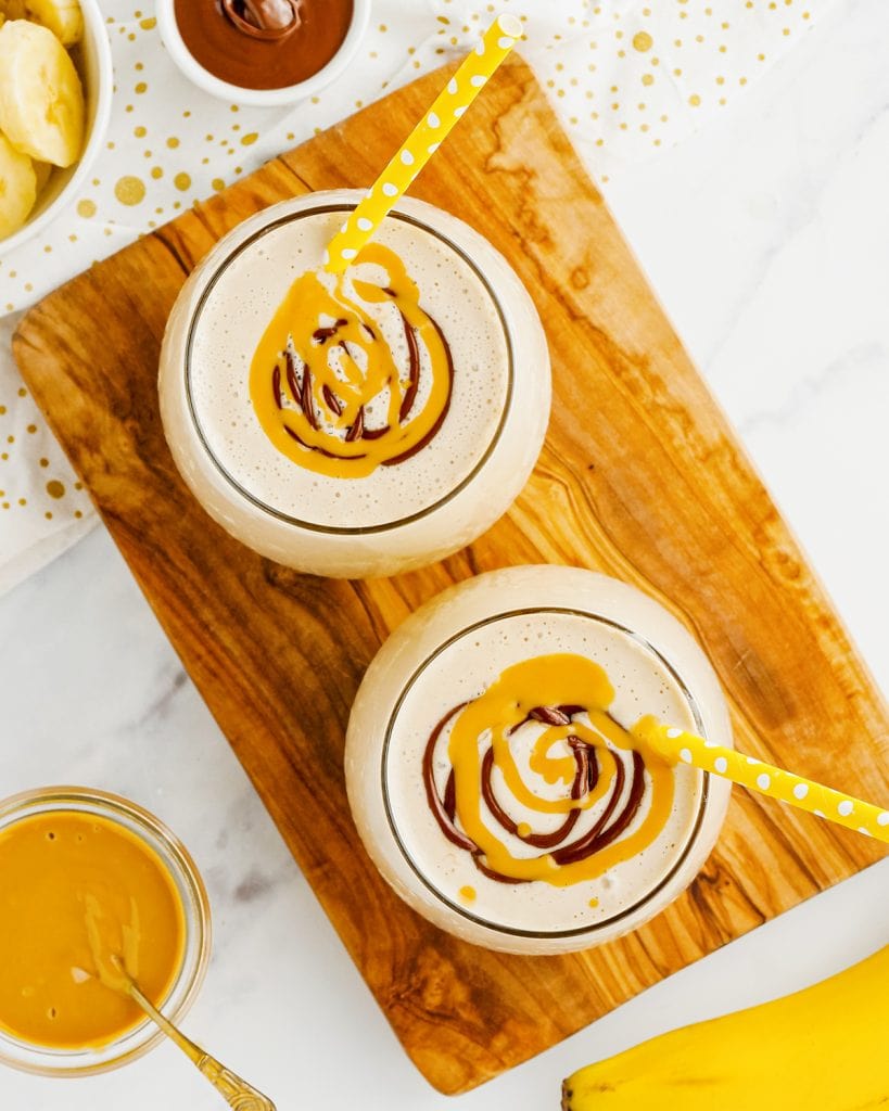 An overhead photo of two glasses of peanut butter smoothie topped with swirls of peanut butter and chocolate, and with yellow straws in the cups.