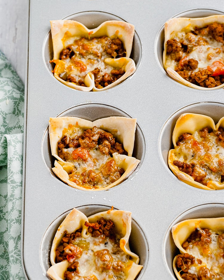 Wonton taco cups in a baking pan, showing a wonton wrapper filled with ground beef, and topped with melted cheese.