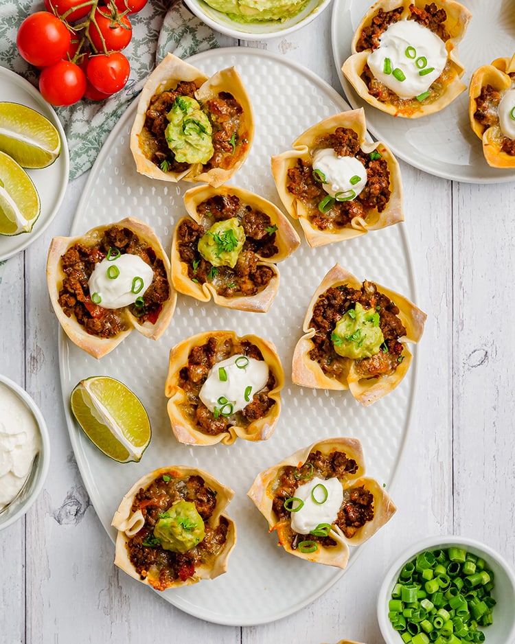 Wonton taco cups on a plate, they are little flower shaped wonton wrappers, filled with ground beef, and each topped with sour cream or guacamole.