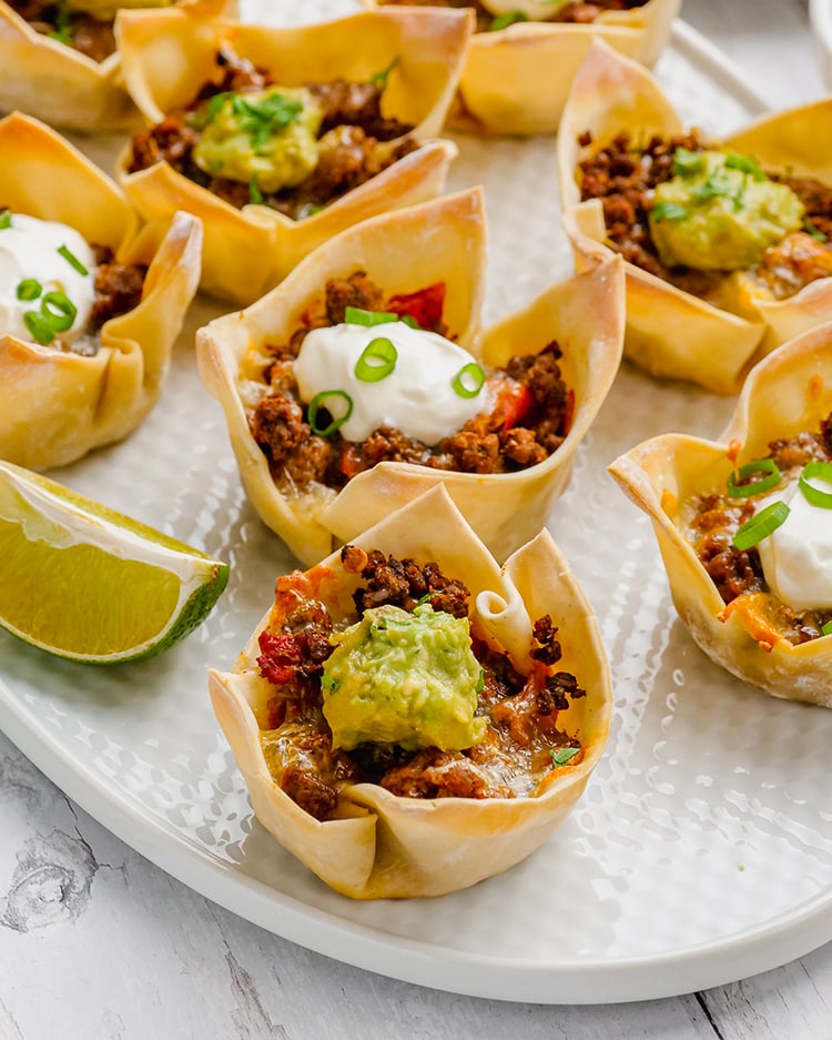 Wonton taco cups on a plate, they are little flower shaped wonton wrappers, filled with ground beef, and each topped with sour cream or guacamole.