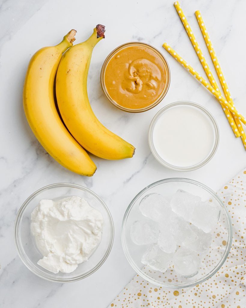 An overhead photo of the ingredients needed to make a peanut butter banana smoothie, bananas, creamy peanut butter, milk, vanilla yogurt, and ice.