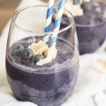 A glass of blueberry smoothie topped with coconut, blueberries, and two straws.