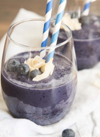 A glass of blueberry smoothie topped with coconut, blueberries, and two straws.