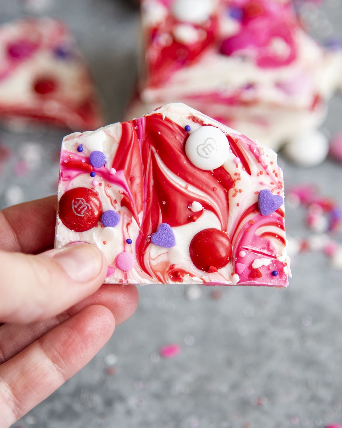 A hand holding a piece of white chocolate bark with swirls of red and pink chocolate and m&ms on top.