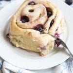 Angled image of blueberry sweet roll on a white plate with a fork cutting in.