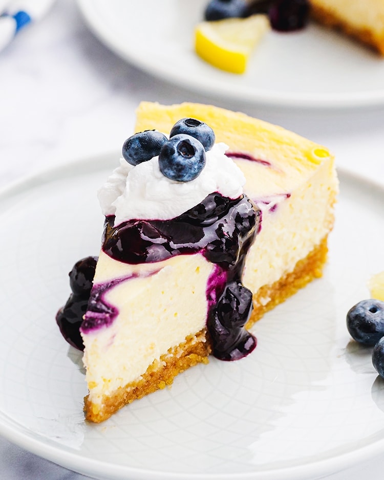 A slice of blueberry cheesecake on a white plate topped with blueberry sauce, whipped cream, and fresh blueberries.