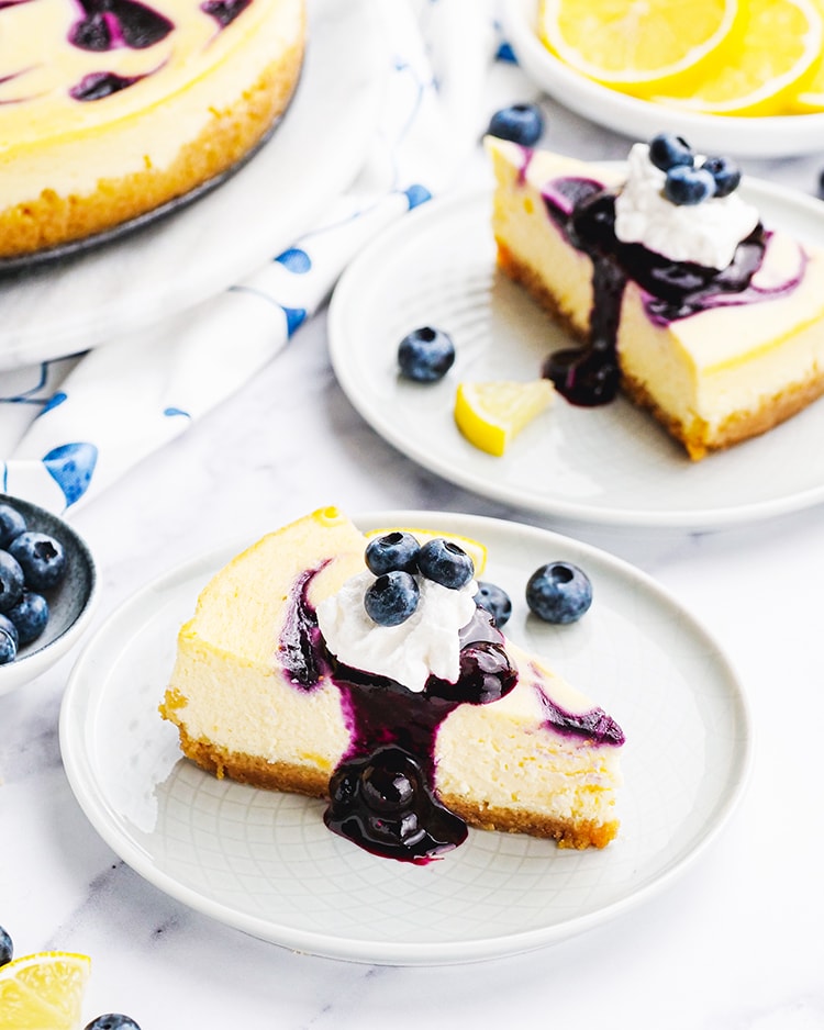 A slice of blueberry cheesecake on a white plate topped with blueberry sauce, whipped cream, and fresh blueberries.