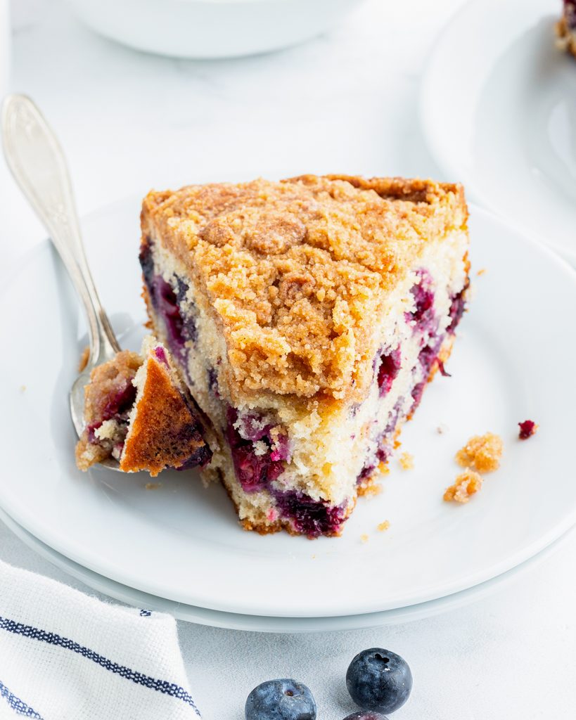 A slice of blueberry coffee cake on a plate with a fork bite out of it.