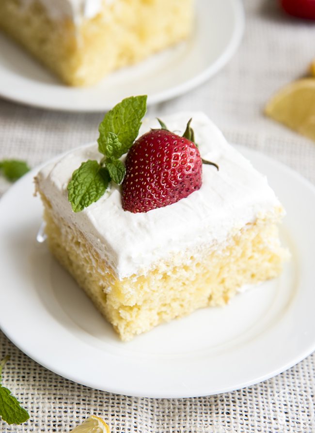 A slice of a yellow cake topped with a white frosting and a half a strawberry and a mint sprig on a small plate.