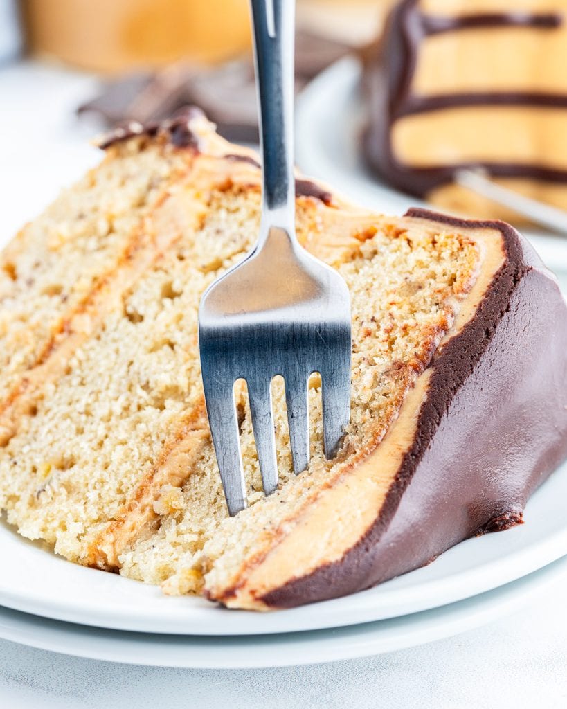A fork sticking into a piece of banana cake topped with banana frosting and chocolate ganache.