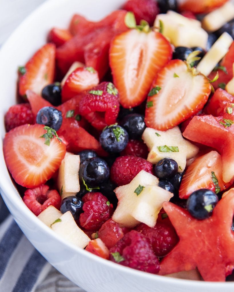 A fruit salad with red, white, and blue fruit in a white bowl, topped with fresh mint pieces.