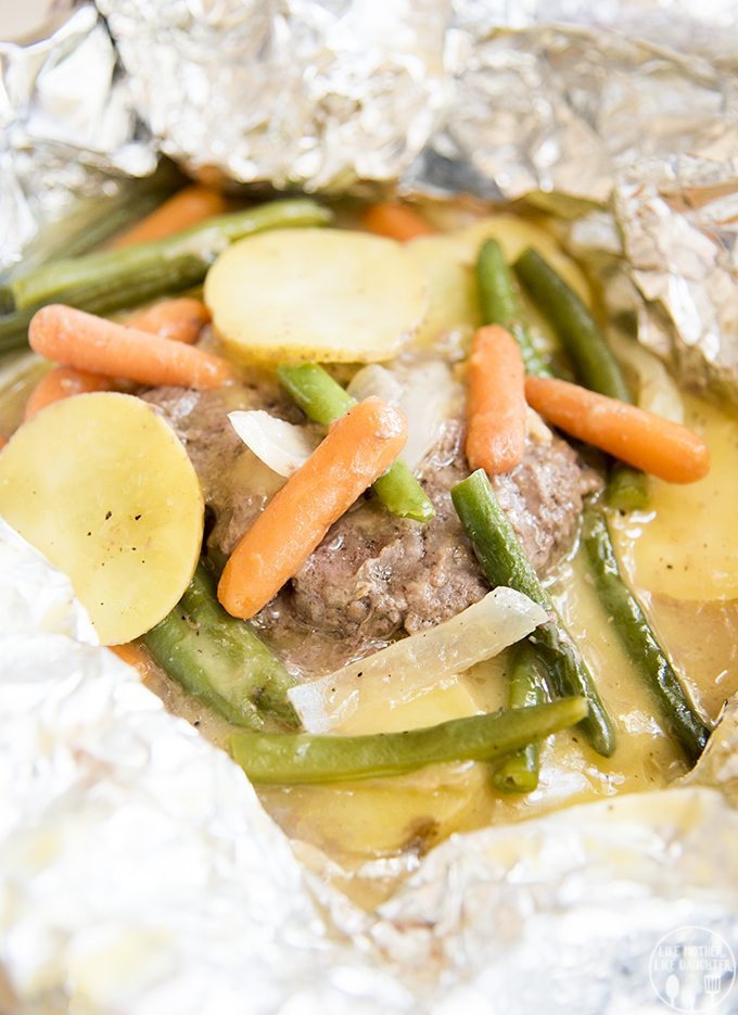 A beef patty covered in green beans, carrots, and potato slices sitting in juices, all wrapped in foil.