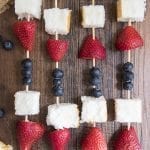 Red, white, and blue fruit kebabs above image on a wood background.