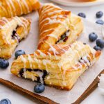 A few blueberry turnovers arranged on a piece of parchment on a wooden tray, with other blueberries scattered around them.