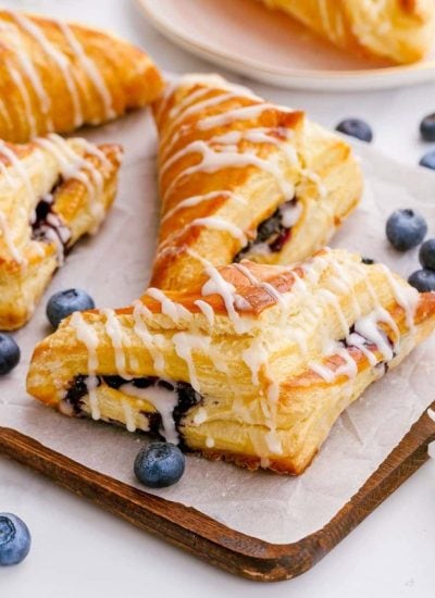 A few blueberry turnovers arranged on a piece of parchment on a wooden tray, with other blueberries scattered around them.