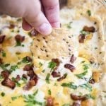 A cheesy dip in a bacon pan, it is topped with bacon pieces, and parsley, and a tortilla chip dipped into it.