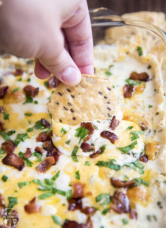 A cheesy dip in a bacon pan, it is topped with bacon pieces, and parsley, and a tortilla chip dipped into it.