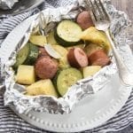 Above image of sausage and veggie foil dinner in foil on a white plate with a fork.