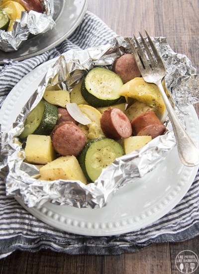 Above image of sausage and veggie foil dinner in foil on a white plate with a fork.