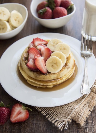 Strawberry banana waffles stacked on a plate with syrup and apples and bananas on top.