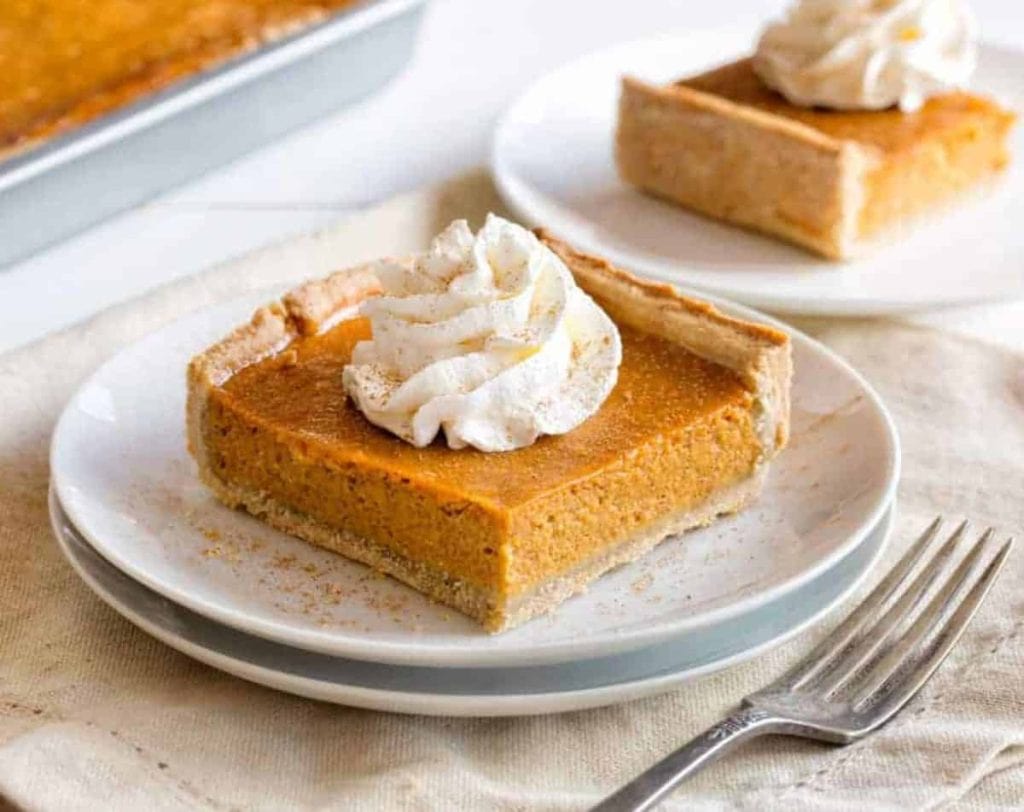 A slice of pumpkin slab pie on a plate topped with whipped cream, and another slice of pie on a plate behind it.