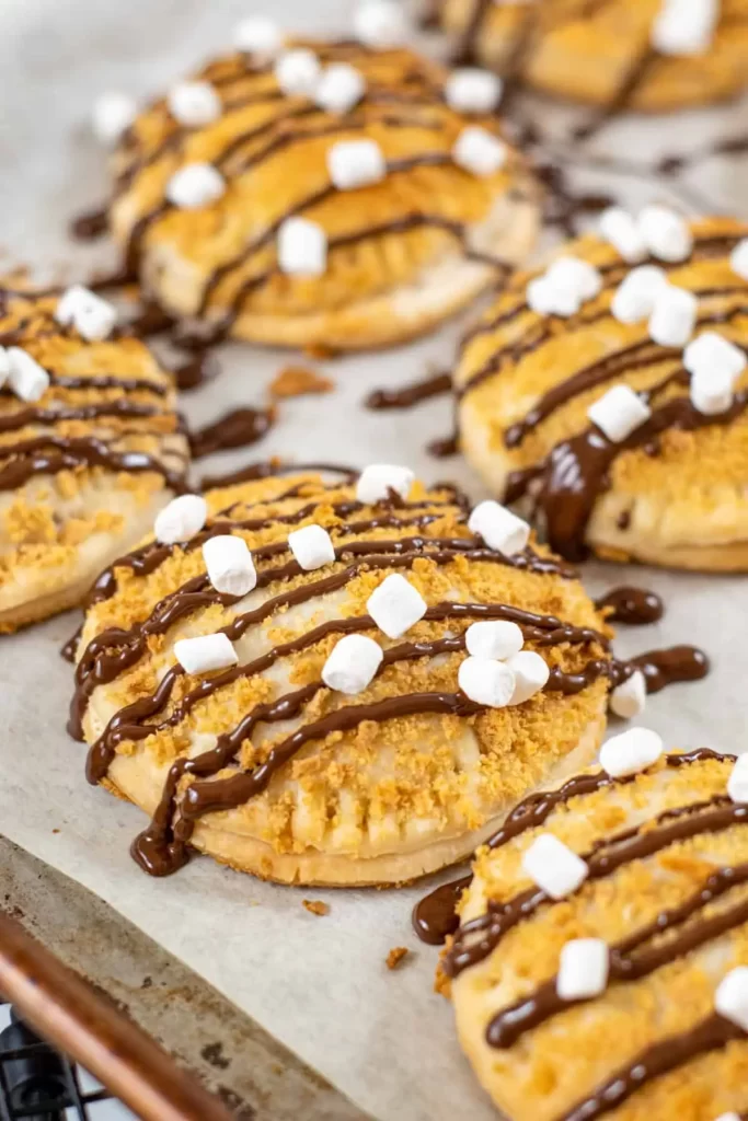 A bunch of circular hand pies on a cookie tray topped with a chocolate drizzle and mini marshmallows. 
