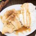 Two apple crescent rolls on a plate with two scoops of vanilla ice cream and caramel sauce over the top.