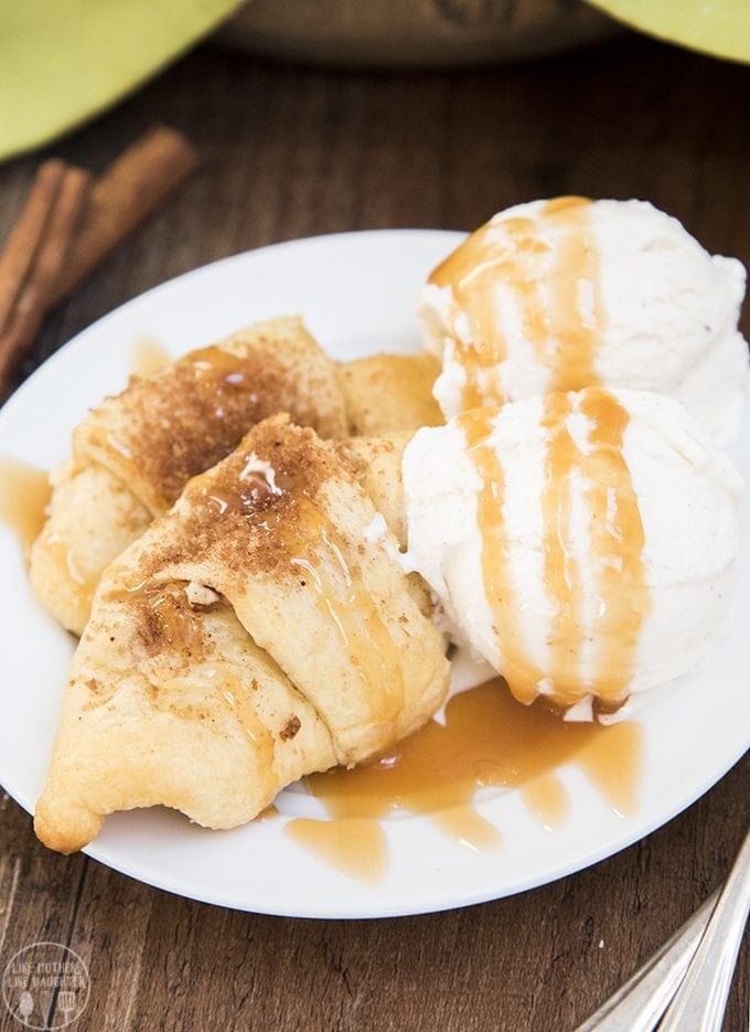 Two apple crescent rolls on a plate with two scoops of vanilla ice cream and caramel sauce over the top.