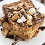A stack of s'mores french toast on a plate topped with graham cracker crumbs, chocolate chips, and mini marshmallows.