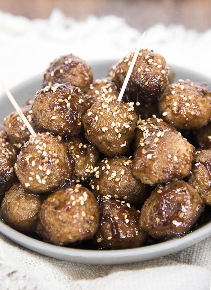 A bowl of teriyaki coated meatballs. Two of the meatballs have toothpicks in them. They are all sprinkled with sesame seeds.