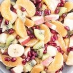 A bowl of fruit salad with pomegranate seeds, oranges, kiwi, banana, and apples.