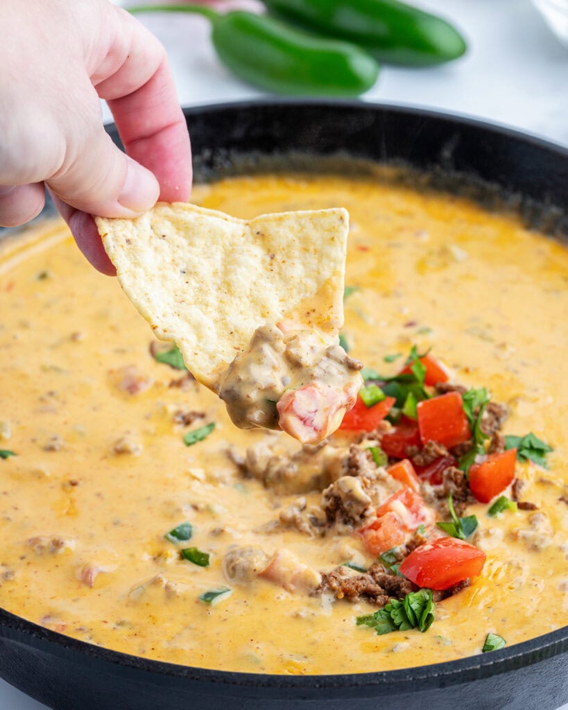 Beef queso dip on a tortilla chip being held by a hand above a skillet of queso dip.