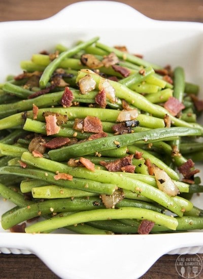A white baking dish full of green beans covered in bacon pieces and sautéed onion.