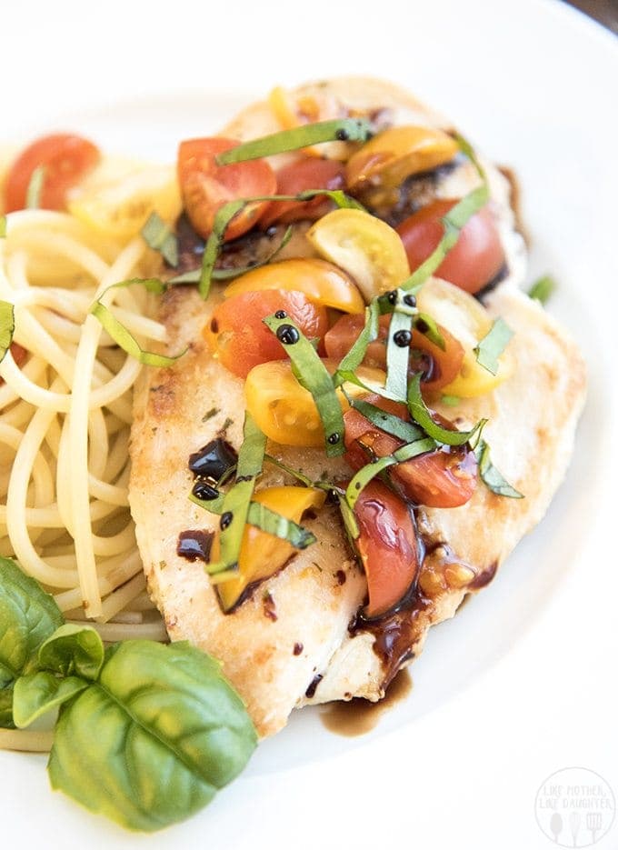 A plate of two chicken breasts topped with cherry tomatoes, fresh basil strips, and drips of balsamic vinegar next to spaghetti noodles.
