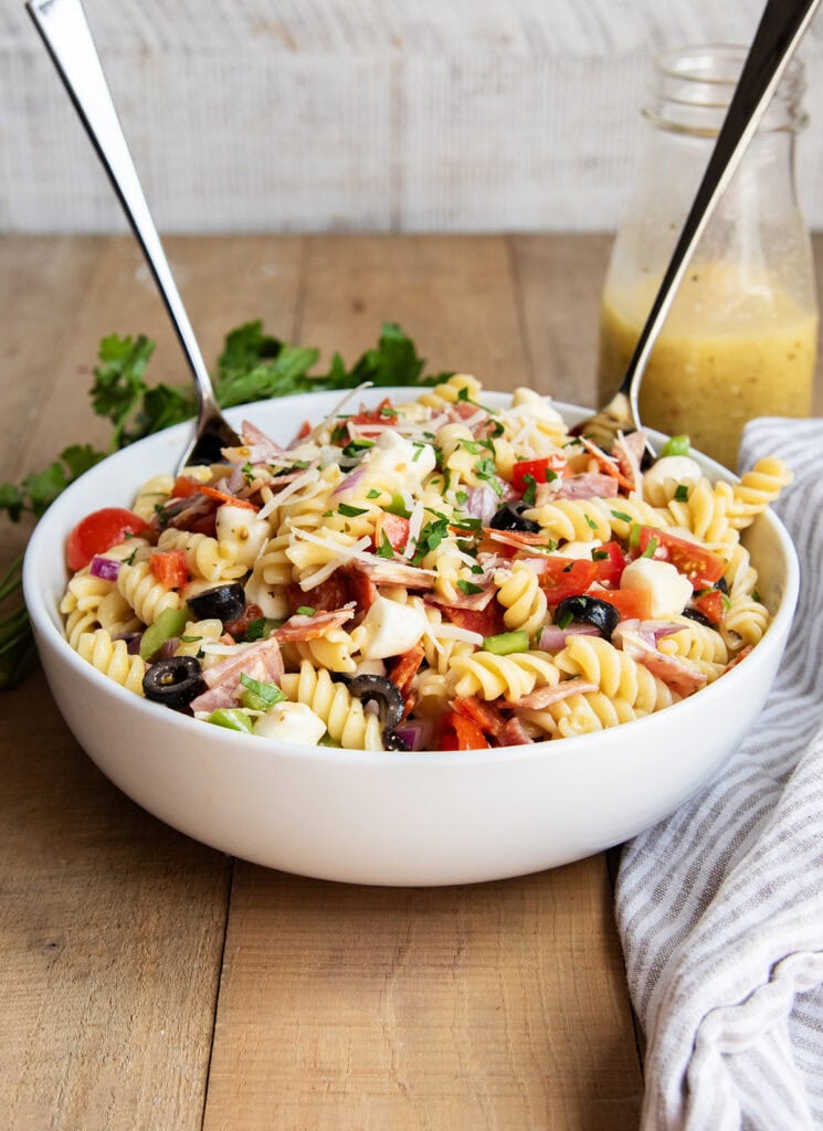 A bowl of pasta salad with two spoons in it, and a bottle of Italian dressing.
