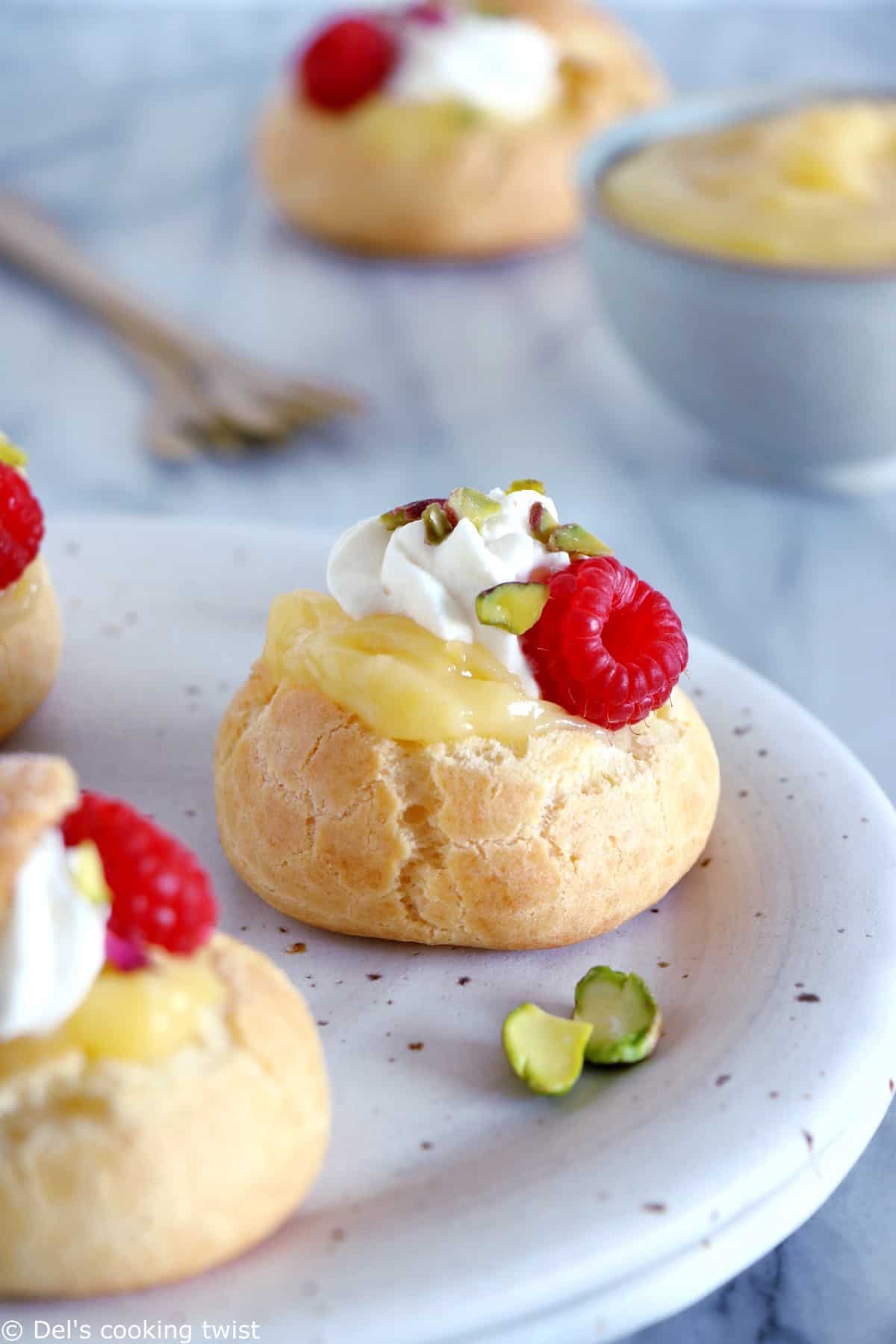 A round cream puff topped with lemon curd, whipped cream, and a fresh raspberry.