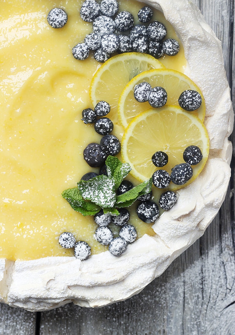 A close up of a white, round pavlova shell topped with lemon curd, and powdered sugared blueberries.