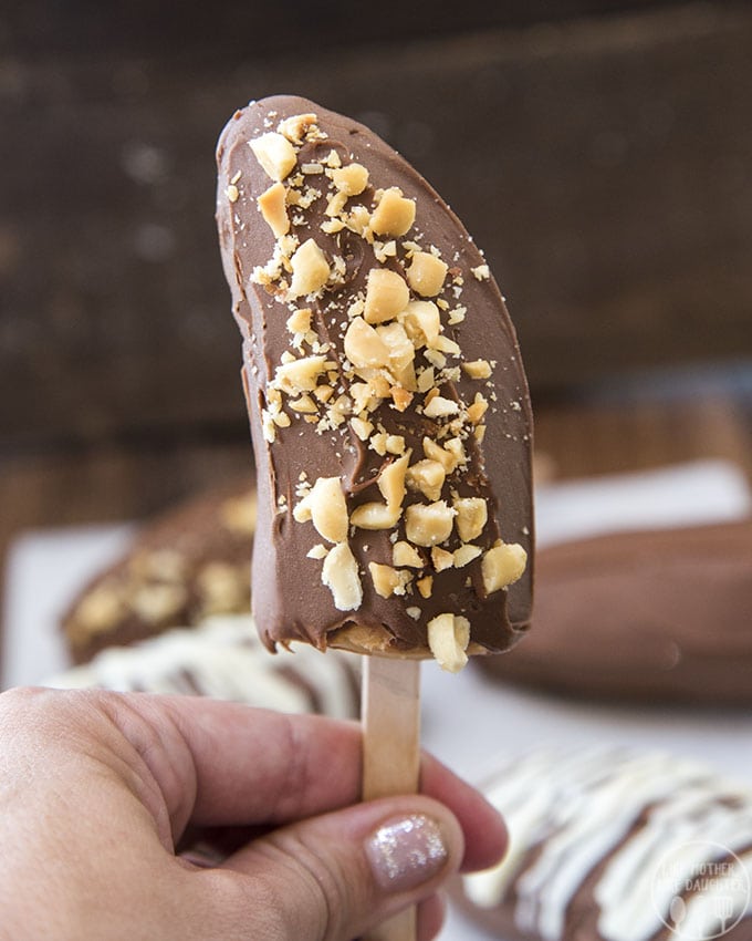A hand holding a chocolate covered banana topped with chopped peanuts. 