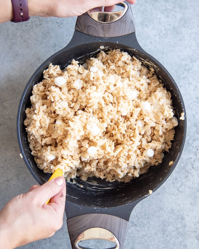 A top view of two hands stirring a pot of rice krispie cereal with marshmallows.