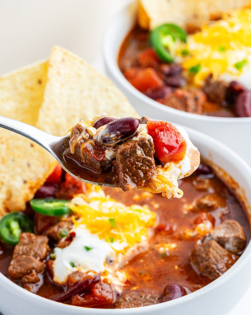 A spoonful of steak chili above a bowl of it.