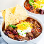 A bowl of steak chili topped with tortilla chips, sour cream, and cheddar cheese.