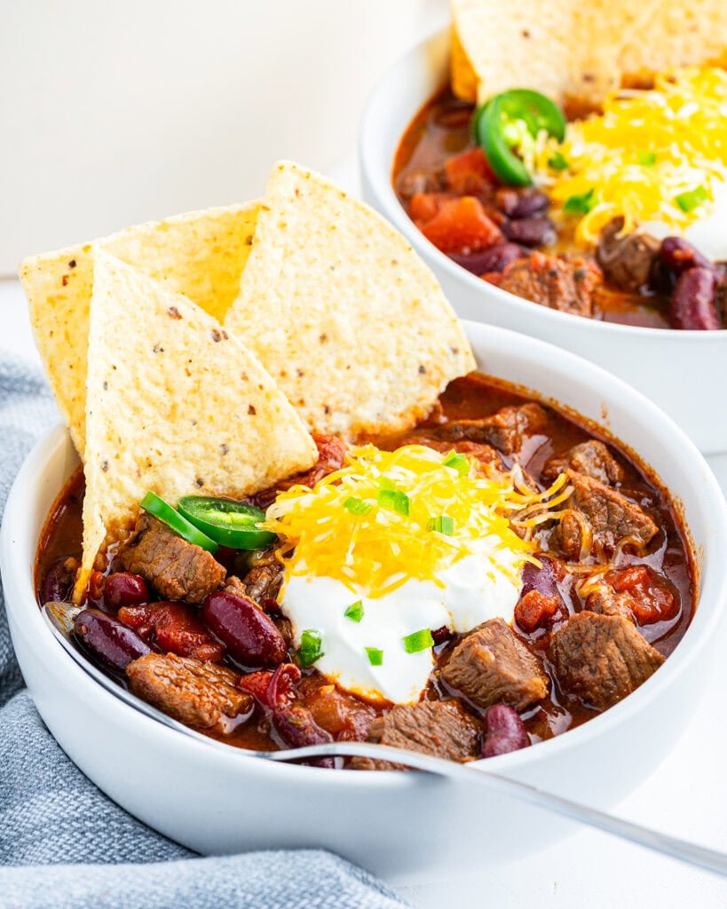 A bowl of steak chili topped with tortilla chips, sour cream, and cheddar cheese.